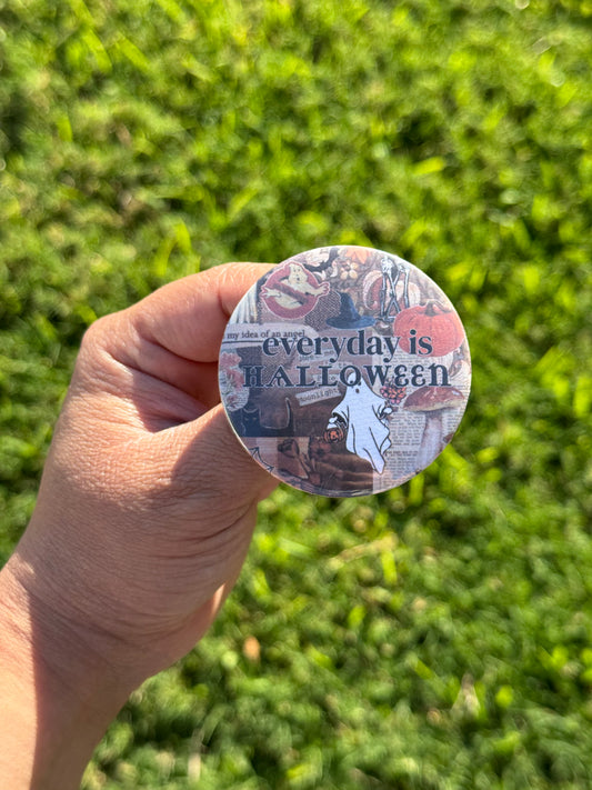 Hand holding a button pin with text that says “Everyday is Halloween.” Background design features image in collage style with various images of ghosts, pumpkins cats and mushrooms 